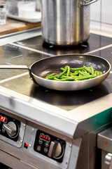 Fresh green beans sautéing in a pan on a modern kitchen stove, with a stainless steel pot in the background, showcasing a vibrant cooking scene.