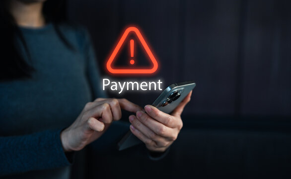 A person is using a smartphone, displaying a warning symbol related to payment issues in a dimly lit environment.