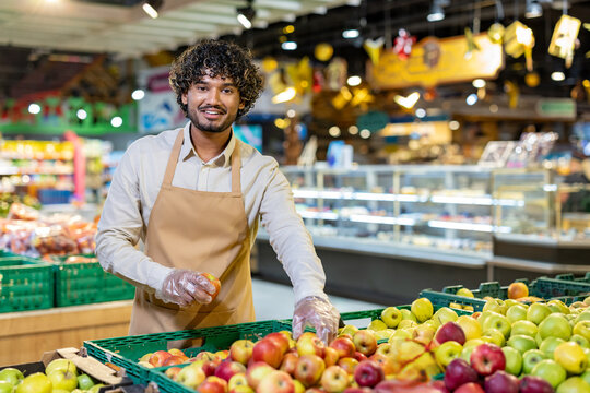 A friendly grocery store employee holds an apple while smiling at the camera, surrounded by fresh produce.