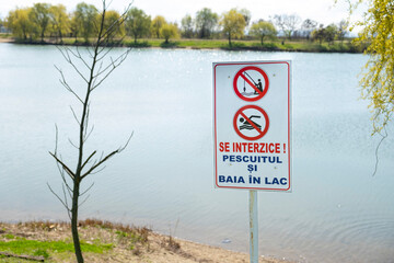 A prohibition sign near a lake warns against fishing and swimming. The sign is in Romanian and stands against a natural background with water and green trees, emphasizing safety regulations in nature.