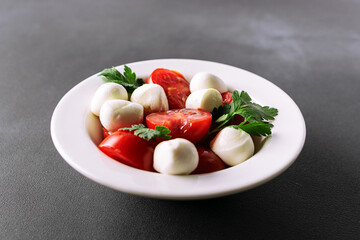 Juicy caprese salad with baby mozzarella cheese, cherry tomatoes and parsley in white ceramic plate on black stone table close up. Healthy diet food concept.