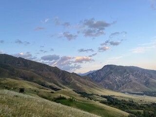serene mountain valley at sunset with rolling green hills, dramatic rocky peaks, and a small village nestled in the landscape

