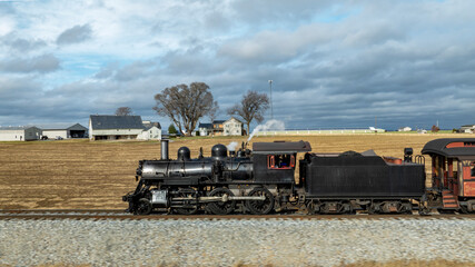 A vintage steam locomotive steams along railway tracks, passing fields and farmhouses under an overcast sky. The scenery reflects a tranquil rural setting.