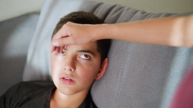 Close-up of someone checking sick boy health by placing hand on his forehead, child slowly opens eyes, showing signs of illness, concern and care in a comforting home environment