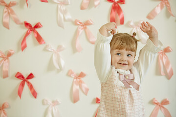 One-and-a-half-year-old girl in a tweed dress and two ponytails held her hands up against a wall of pink bows