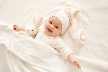 a happy newborn baby before going to bed in a sleeping cap under a blanket lies on the back and smiles on the white bed in the bedroom, a happy cute little baby, top view