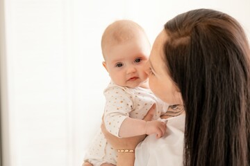 A mother gently kisses and hugs a newborn baby holding him in her arms in a bright bedroom,...