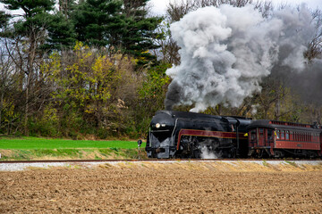 A vintage steam locomotive travels along countryside tracks, releasing clouds of black smoke into the crisp autumn air. Colorful trees provide a backdrop as the train moves forward, evoking nostalgia.