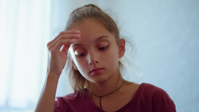 Close up of little girl with ponytail and maroon shirt resting head on hand while looking down with sober thoughtful expression in soft indoor light showing emotion