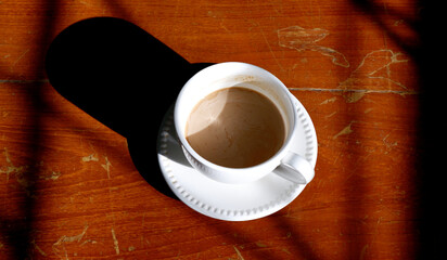 Hot coffee in white cup placed on vintage wooden table with strong sunlight in morning. Coffee cup latte texture top view. Black shadow, breakfast, energy, wood, rustic kitchen, cafe and drink concept