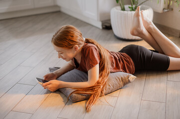 A red-haired girl with long hair is lying on the kitchen floor playing and scrolling her phone....