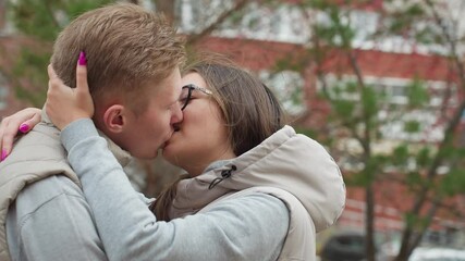Two lovers share passionate kiss outdoors in matching outfits, wind gently sways hair and autumn leaves around them as they embrace closely