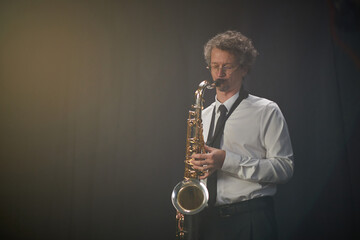 Adult male saxophonist wearing white shirt and tie playing saxophone with passion standing against...