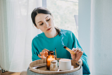 Young woman burning palo santo near a tray with candles and tea in a calm home setting. Sensory...