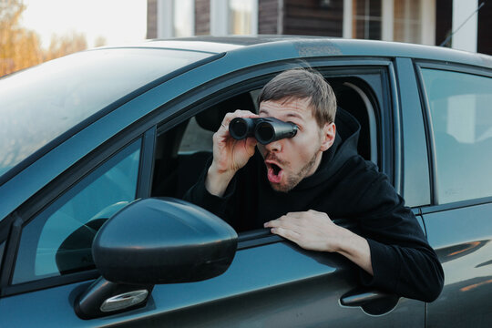 Young shocked man stalker watching through binoculars sitting in car. Man spying or investigating.