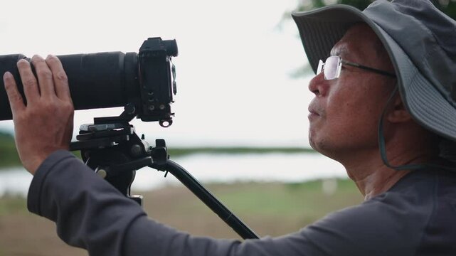 profile of asian photographer with big hat adjusting tripod and zoom of reflex camera
