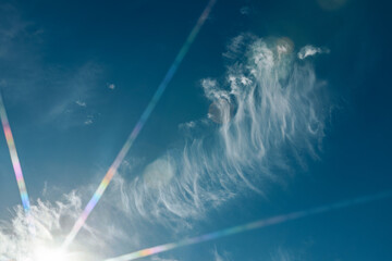 Thin, streaky clouds drift across a deep blue sky as sunlight creates colorful lens flares, adding...