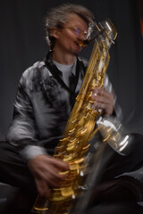 Vertical dynamic shot of male musician expressively playing saxophone sitting in grey studio with...