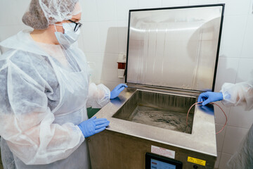 Female technician in sterile clothing examining warm water bath inside breast milk pasteurizer. Equipment preparation before donor milk processing cycle.