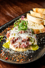 A gourmet dish featuring finely chopped meat topped with grated cheese and herbs, served alongside slices of crusty bread on a dark plate, with a sprinkle of spices.