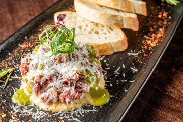 A gourmet dish featuring finely chopped meat topped with grated cheese and herbs, served alongside slices of crusty bread on a dark plate, with a sprinkle of spices.