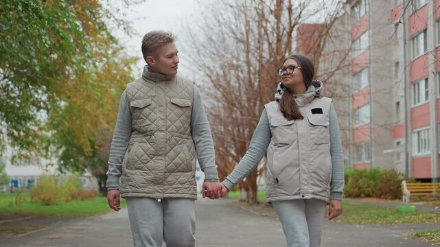 Joyful moment of young couple walking hand in hand, swinging arms while smiling and looking at each other with affection during peaceful stroll along tree-lined pathway with blurred urban background