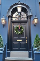An elegant black door, beautifully decorated with a festive wreath and flanked by warm, glowing lanterns, stands out in a charming urban setting during the joyous holiday season