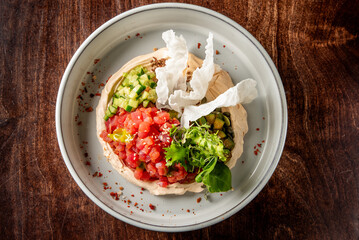 A beautifully arranged plate featuring creamy hummus topped with fresh tomato, cucumber, and herbs, garnished with crispy crackers, all set against a rustic wooden background.