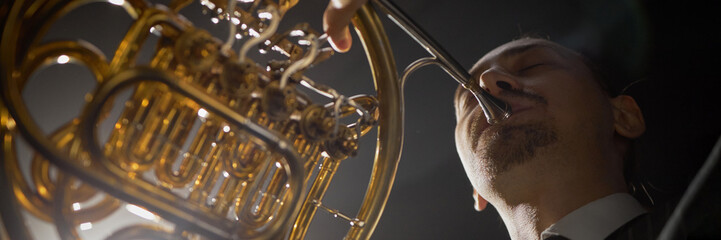 Website header shot concentrated male musician with eyes closed playing French horn creating...