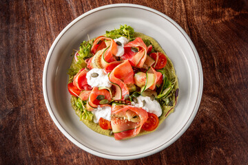 A vibrant salad featuring thinly sliced vegetables, including tomatoes and greens, drizzled with creamy dressing and garnished with seeds, presented in a white bowl on a wooden surface.

