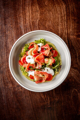 A vibrant salad featuring thinly sliced vegetables, including tomatoes and greens, drizzled with creamy dressing and garnished with seeds, presented in a white bowl on a wooden surface.
