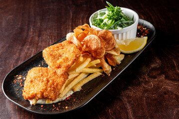 A tempting plate featuring crispy fried fish fillets with golden fries and potato chips, accompanied by a refreshing side of green salad and a lemon wedge, all on a sleek black plate.