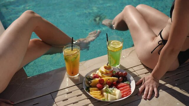 On this sunny day, two women enjoy poolside with colorful cocktails and a platter of fresh exotic fruits
