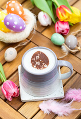 A cup of hot chocolate  toped with mousse of milk, latte art in an Easter egg shape with festive Easter decoration such eggs, colorful tulips and feathers.