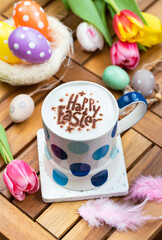 A cup of hot chocolate  toped with mousse of milk, latte art in text "Happy Easter" with festive Easter decoration such eggs, colorful tulips and feathers.