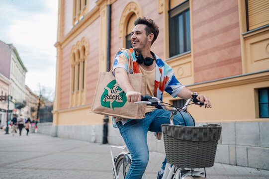 Young man riding bicycle carrying reusable shopping bag promoting sustainability in urban setting - Powered by Adobe