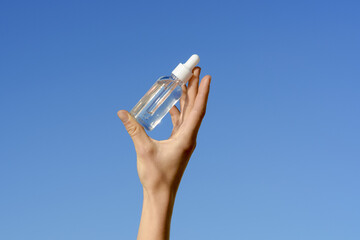 Female hand holding a transparent glass jar of serum with a pipette dispenser on a blue sky...