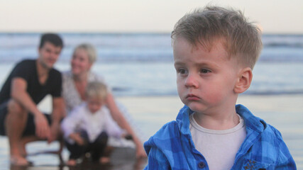 closeup portrait of sad serious toddler contemplating and  his cheerful parents in background on...