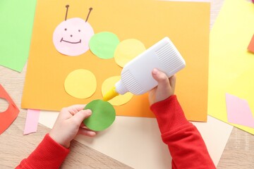 Little girl making handmade project with color paper and glue at wooden table, top view