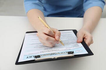 Doctor with pen filling out health insurance policy at white table in clinic, closeup