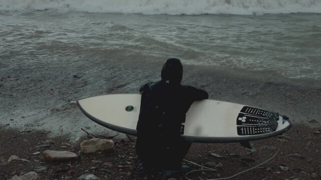 Male surfer in wetsuit stands on the shore, holding surfboard. Man on stone beach in winter in northern ocean or sea, ice surfing. BIg wave and grey sky after snow, cold water.