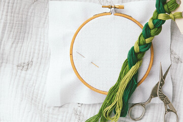 Embroidery hoop with white fabric, two needles, green thread braid and scissors on soft textile,...