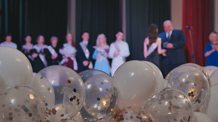 White and transparent balloons filled with gold and silver confetti celebrate graduation, surrounded by blurred friends and teachers