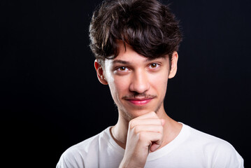 Handsome young man wearing white shirt touching his chin with one hand. Isolated on black...