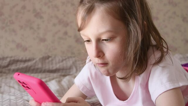 Little girl in pink shirt lies on bed and looks at smartphone with serious focus, absorbed in screen in cozy home atmosphere