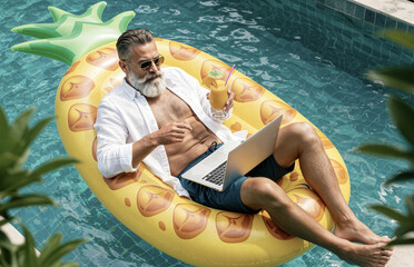 A mature stylish man working remotely on a laptop while lounging on a pool float, embodying productivity, leisure, and relaxation under a sunny outdoor setting