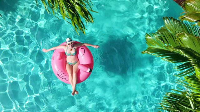 Tropical paradise concept. Ideal summer vacation background with copy space. Aerial view of woman on Pink flamingo ring in blue pool under lush palm trees. Luxury vacation villa in Miami, Florida 4K