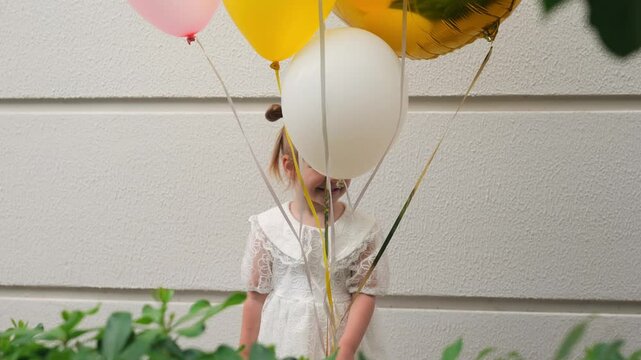 Little girl celebrates her third birthday with colorful balloons near a white wall