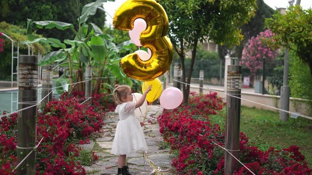 Child celebrates third birthday with colorful balloons in a garden setting filled with flowers