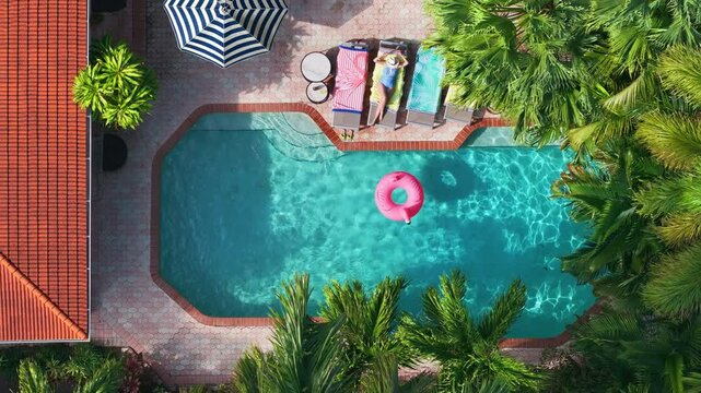 Top view shot of woman relaxing on lounge chair while inflatable pink flamingo floating in light blue water. Aerial view of swimming pool with blue ripples water surface. Holidays recreation concept